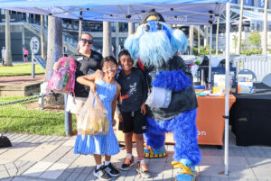 Two children, an adult, and the Tampa Bay Rays mascot standing together holding bags of donated school supplies.
